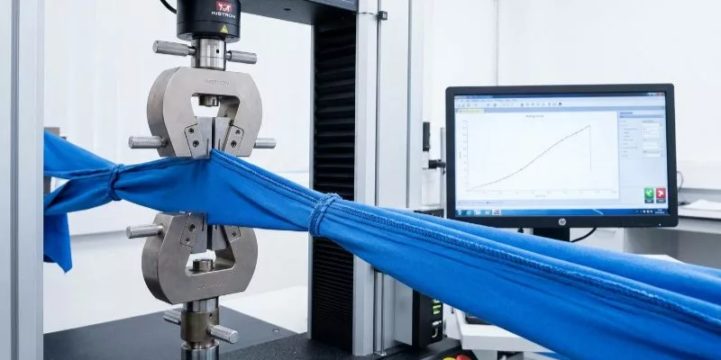 Industrial fabric tensile strength testing machine aggressively pulling apart a sewn seam during an ASTM D1683 lab test, with digital force readings visible.