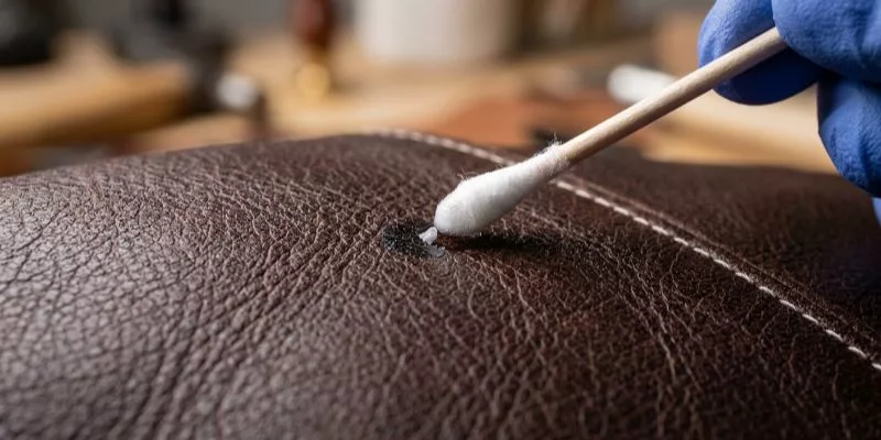 A close-up action shot of a soft horsehair brush gently sweeping away dust from the surface of a leather wallet, following the grain of the leather.